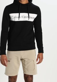 Felpa nera con il logo bianco "Calvin Klein Jeans" sul davanti, dotata di cappuccio con cordino. Abbinata a pantaloni corti beige strappati, con dettagli in patch.