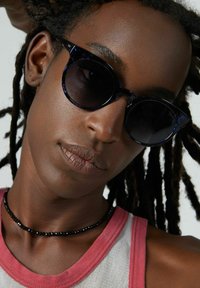 Lunettes de soleil noires rondes avec une monture en bleu marine translucide, portées par une personne aux cheveux bouclés. Un collier de perles noires et un débardeur rose sont visibles.