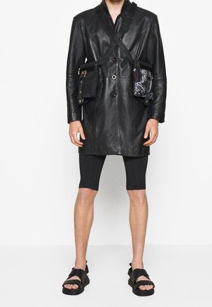 Persona con un abrigo de cuero negro con dos bolsos de bandolera adjuntos, pantalones cortos negros hasta la rodilla y sandalias negras con tiras.