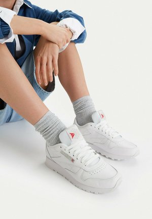 Persona sentada con las manos cruzadas, vistiendo una chaqueta azul remangada, pantalones cortos de mezclilla, calcetines grises y zapatillas blancas Reebok sobre un fondo blanco.