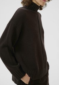 Pull en maille marron foncé avec un col côtelé haut, coupe décontractée et ourlet côtelé. Présente une fente sur le côté et des détails de couture visibles.