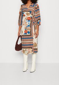 Wikkeljurk in een bloemen- en geometrisch patroon met blauwe, oranje en crèmekleuren. Heeft driekwart mouwen en een strik om de taille, gecombineerd met witte laarzen.