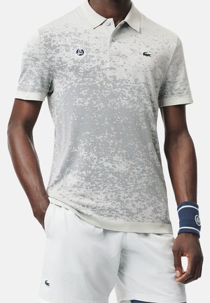 Mand iført hvid og grå mønstret poloshirt med Roland Garros- og Lacoste-logoer, hvide shorts og et blåt armbånd.
