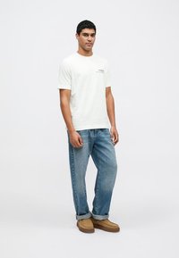 Biała bawełniana koszulka z krótkimi rękawami i małym nadrukiem graficznym, zestawiona z jasnoniebieskimi zwężanymi dżinsami i beżowymi butami wsuwanymi.