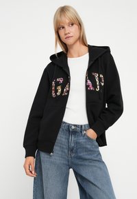 Il felpa nera presenta una scritta floreale "GAP", una chiusura lampo anteriore, un cappuccio con coulisse e polsini a coste. Abbinato a un paio di jeans blu a vita alta.