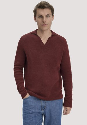 Junger Mann trägt einen dunkelroten, langärmeligen, gestrickten Pullover mit V-Ausschnitt und blaue Jeans, steht mit einer Hand in der Tasche.