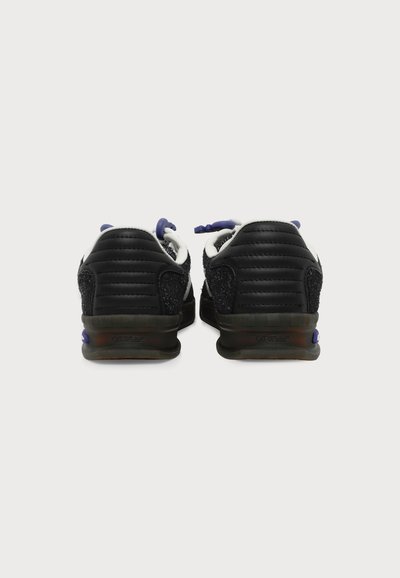 Paire de baskets noires et blanches vues de dos, présentant des matériaux texturés et des lacets violets sur fond blanc uni.