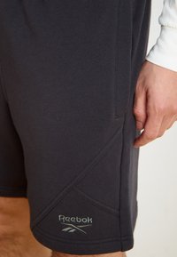 Reebok sporta šorti tumši pelēkā krāsā. Iezīmējas ar gludu audumu, brīvu piegriezumu, izteiktu šuvju dizainu un izšūtu logotipu kreisajā apakšējā kājā.