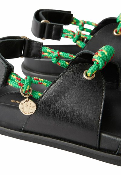 Sandalias de cuero negro con tiras trenzadas de cuerda en verde, amarillo y rojo, y un pequeño charm circular dorado adjunto.