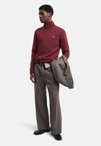 Maglione a dolcevita bordeaux, pantaloni a quadri grigi e scarpe nere. Il modello tiene in mano un blazer grigio; la camicia a righe è visibile all'altezza della vita.