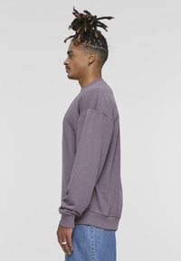 Felpa a maniche lunghe con collo rotondo in tessuto viola tenue, con polsini e orlo a coste; indossata con jeans in denim azzurro chiaro.