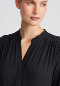 Schwarze Bluse mit Stehkragen, gerafften Details an den Schultern und tiefem V-Ausschnitt. Glatter Stoff mit weicher Textur.