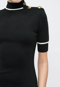 Μαύρο ριμπ μπλουζάκι με κοντά μανίκια και λευκή επένδυση στο γιακά και τα μανίκια, με τρία χρυσά κουμπιά στον δεξί ώμο.