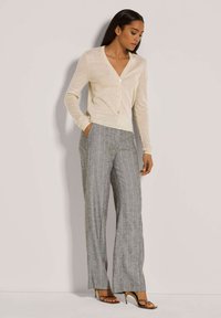 Frau, die eine beigen, knopfgeschlossenen Strickjacke, graue, gestreifte weite Hosen und braune hohe Sandalen trägt, steht da mit einer Hand in der Tasche.