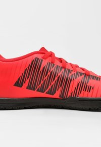 Zapatillas deportivas rojas con una parte superior sintética suave, que presenta rayas diagonales negras y el logo de la marca. Suela de goma con un patrón de tracción texturizado.