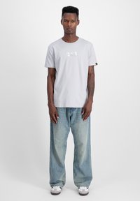 Gai pelēks īsām piedurknēm T-krekls ar metāliski taisnstūrveida logotipu uz krūtīm, apvienots ar brīvi piegulošiem vidēji ziliem džinsiem.