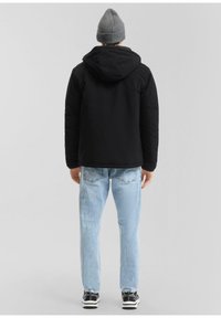 Zwarte gewatteerde jas met capuchon, lichtblauwe jeans en zwarte sneakers. Model draagt een grijze gebreide muts, gezien van achteren.