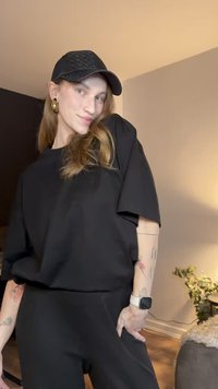 Fekete, bő szabású rövid ujjú felső fekete nadrággal párosítva. Fekete, texturált sapkát és arany karika fülbevalókat visel. Fehér okosóra a csuklój�án.
