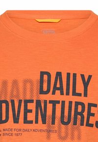 Pomarańczowa koszulka z czarnym napisem "DAILY ADVENTURES" oraz mniejszym tekstem poniżej: "MADE FOR DAILY ADVENTURES SINCE 1977."