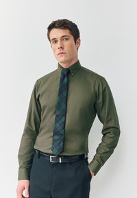 Camisa verde de botones con cuello, combinada con una corbata oscura de cuadros en verde y azul marino. Se lleva con pantalones negros y cinturón.