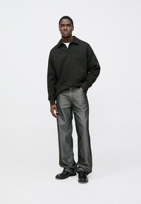 Fekete gyapjú pulóver gallérral és mellkason lévő logóval, szürke, bő szárú farmerrel és fekete cipővel, egyszerű háttér előtt állva.