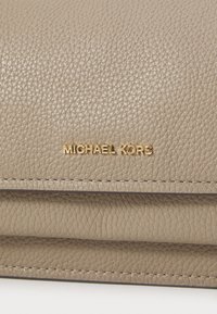 Bēša ādas soma ar akmens faktūru; ar redzamām šuvēm un zelta "MICHAEL KORS" logotipu izsistā formā priekšpusē.