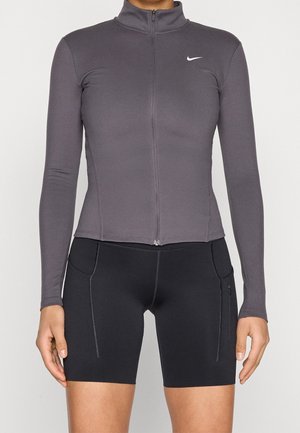 Persona che indossa una maglia grigia aderente a maniche lunghe con zip e pantaloncini da ciclismo neri a vita alta con cuciture visibili e tasche.