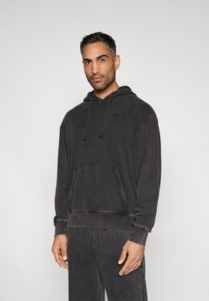 Άνδρας που φοράει ένα ξεθωριασμένο μαύρο φούτερ με κουκούλα και μπροστινή τσέπη, και μαύρο παντελόνι που ταιριάζει, στέκεται μπροστά σε ένα απλό φωτεινό φόντο.