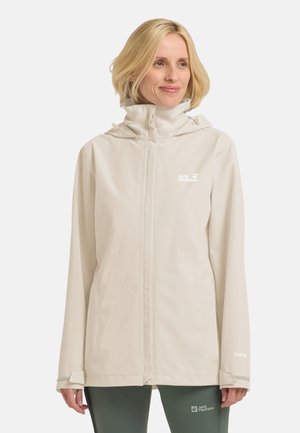 Donna che indossa una giacca con cappuccio Jack Wolfskin beige chiaro e pantaloni Jack Wolfskin verde scuro, sorridente e che guarda leggermente a destra.