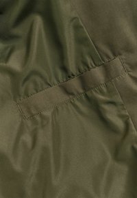 Green nylon fabric with a smooth texture, featuring a horizontal pocket with a flat closure and subtle sheen, primarily in an olive tone.