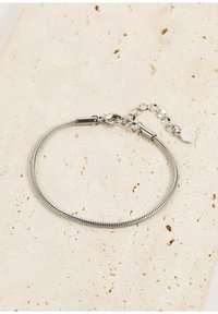 Pulsera de cadena de serpiente plateada con un cierre de langosta, longitud ajustable y un charm circular que presenta un patrón texturizado. Colocada sobre una superficie de piedra clara.