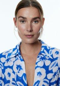 Hemd mit blau-weißem Blumenmuster, ausgestattet mit einem umgeschlagenen Kragen und Knopfverschluss. Leichter Stoff mit halbtransparenter Textur.