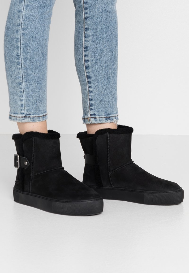 Bottines en daim noir avec une tige douce et texturée, ornées d'une boucle et d'une semelle en caoutchouc plate, associées à un jean bleu raccourci.