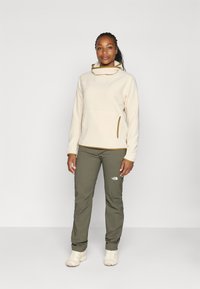 Crème fleece pullover met hoge kraag, contrasterende bruine accenten en ritsen. Olijfgroene broek met een logo op de dij. Lichte schoenen.