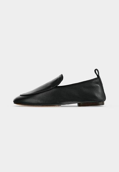 Μαύρο δερμάτινο παπούτσι τύπου slip-on με στρογγυλεμένο νύχι, χαμηλό τακούνι και πίσω λουράκι, σε μονόχρωμο ανοιχτό φόντο.