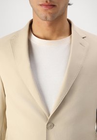 Béžový blazer s přiléhavým střihem, na jediné knoflíkové zapínání a s klopami. Nositelný přes bílé tričko, s hladkou texturou látky.
