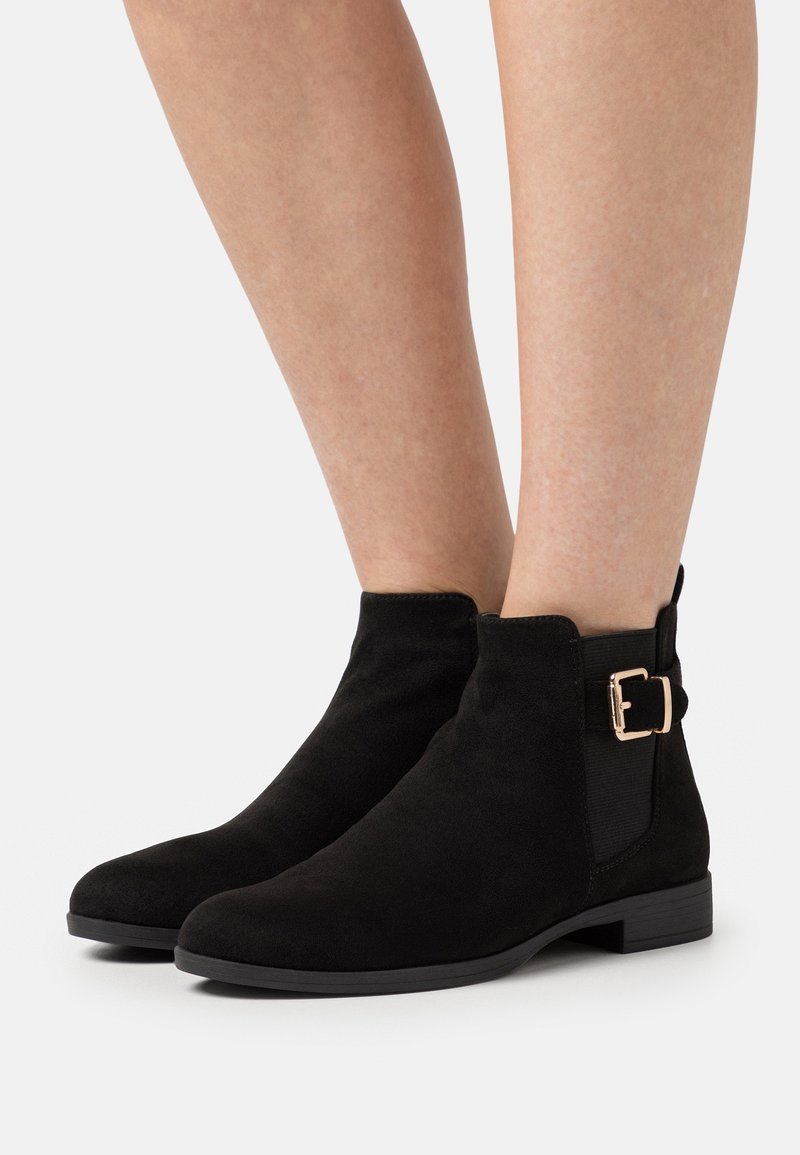 Bottes basses en daim noir avec talons bas, panneaux élastiques latéraux et brides décoratives à boucle dorée, portées sur des jambes nues.