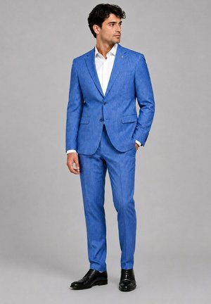 Homme portant un costume bleu vif ajusté avec une chemise blanche et des chaussures noires, debout avec une main dans sa poche sur un fond gris.