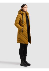 Femme debout portant un manteau matelassé à capuche jaune moutarde, des leggings noirs et des bottines noires sur fond gris.