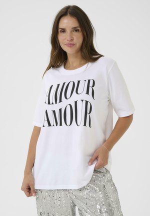 Hvid bomuld t-shirt med "AMOUR" trykt i fed sorte bogstaver. Korte ærmer, afslappet pasform. Model iført sølv palietterbukser.