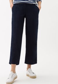 Pantalon en lin bleu marine à jambes larges avec poches latérales, associé à des espadrilles blanches à lacets. Texture lisse, design décontracté.