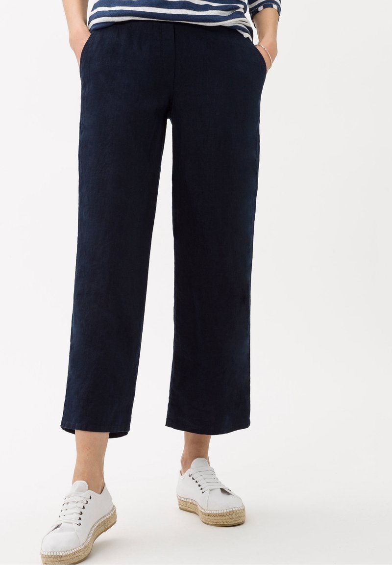 Pantalon en lin bleu marine à jambes larges avec poches latérales, associé à des espadrilles blanches à lacets. Texture lisse, design décontracté.