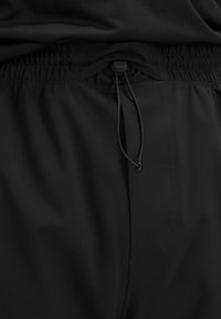 Detailný záber čiernych elastických nohavíc s nastaviteľným šnúrkovým pásom a malým logom na svižnom uzávere.