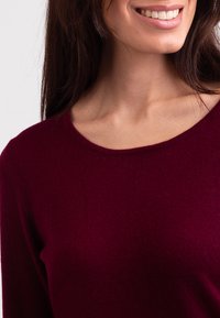 Burgundowy sweter dzianinowy z okrągłym dekoltem, o miękkiej teksturze i dopasowanym kroju, koncentrując się na kołnierzu i górnej części odzieży.