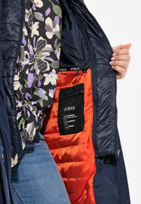 Giacca impermeabile navy con fodera arancione, tasche con zip e etichetta interna nera. Camicia floreale visibile sotto.