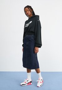 Sweatshirt preto com capuz e logótipo branco, saia de ganga de cintura alta, meias brancas e ténis coloridos. Modelo em p�é sobre um fundo azul.