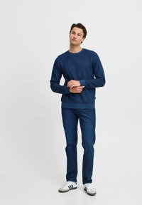 Felpa blu navy con collo rotondo, polsini e orlo a costine, abbinata a jeans di denim scuro e sneaker bianche con strisce nere.