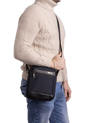 Sac noir en nylon à bandoulière avec une surface texturée, poche avant zippée et accents en cuir, porté à la hanche avec une sangle réglable.