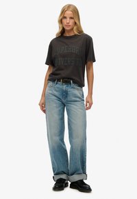 Camiseta gris oscuro con la impresión "Superdry University", combinada con jeans rectos de mezclilla azul claro. Jeans enrollados en los puños, zapatos negros.