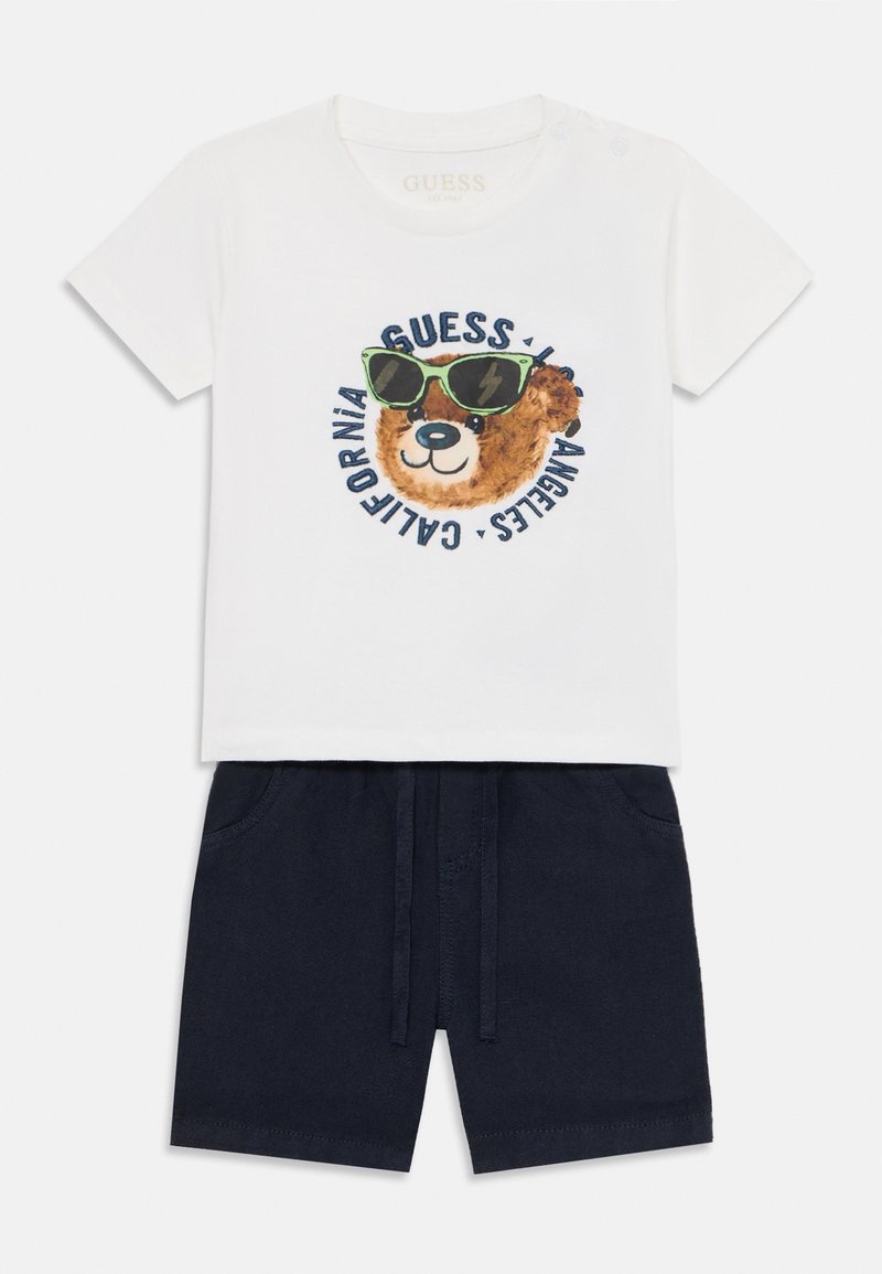 Witte katoenen t-shirt met een berenafbeelding die groene zonnebril draagt, gecombineerd met marineblauwe shorts met een elastische band en zijzakken.